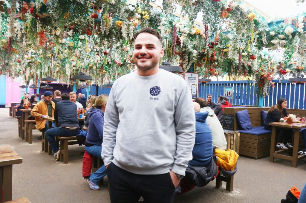 A man in a grey sweatshirt with the Blue Collar street food bar behind him. There are hanging gardens 