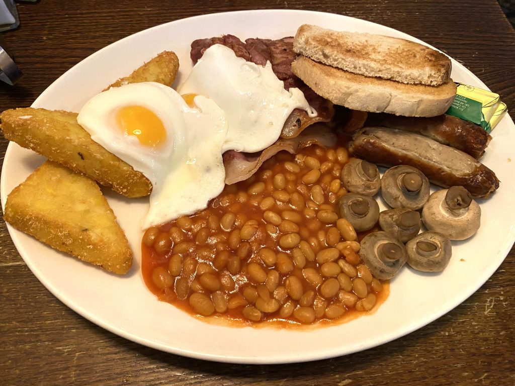 A plate of hash browns, fried eggs, bacon, toast and butter, sausages, mushrooms and baked beans