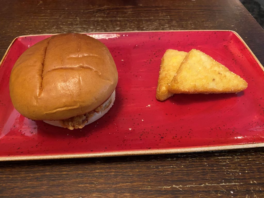 The Cornflake Chicken Burger with two hash browns on a red, rectangular plate