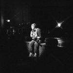 Benjamin Franics Leftwich performing on a stage. He is seated, playing a guitar while a spotlight focuses on him