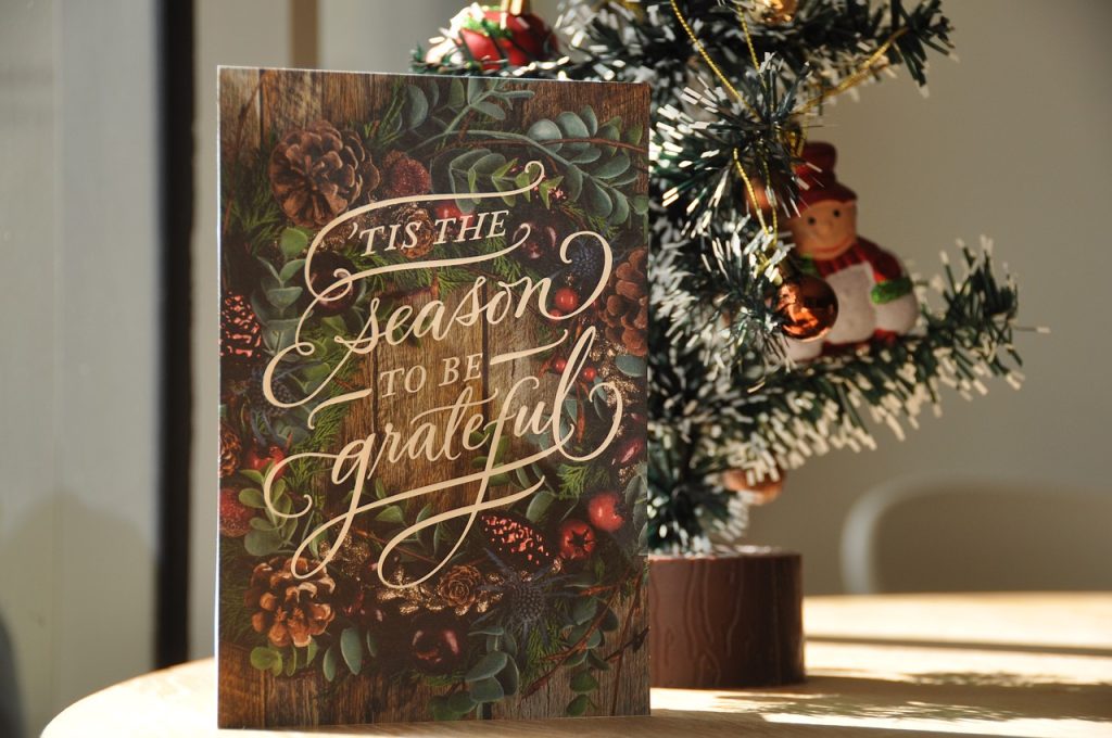 A Christmas card on display with a tree behind it. The card's text reads 'Tis The Season To Be Grateful