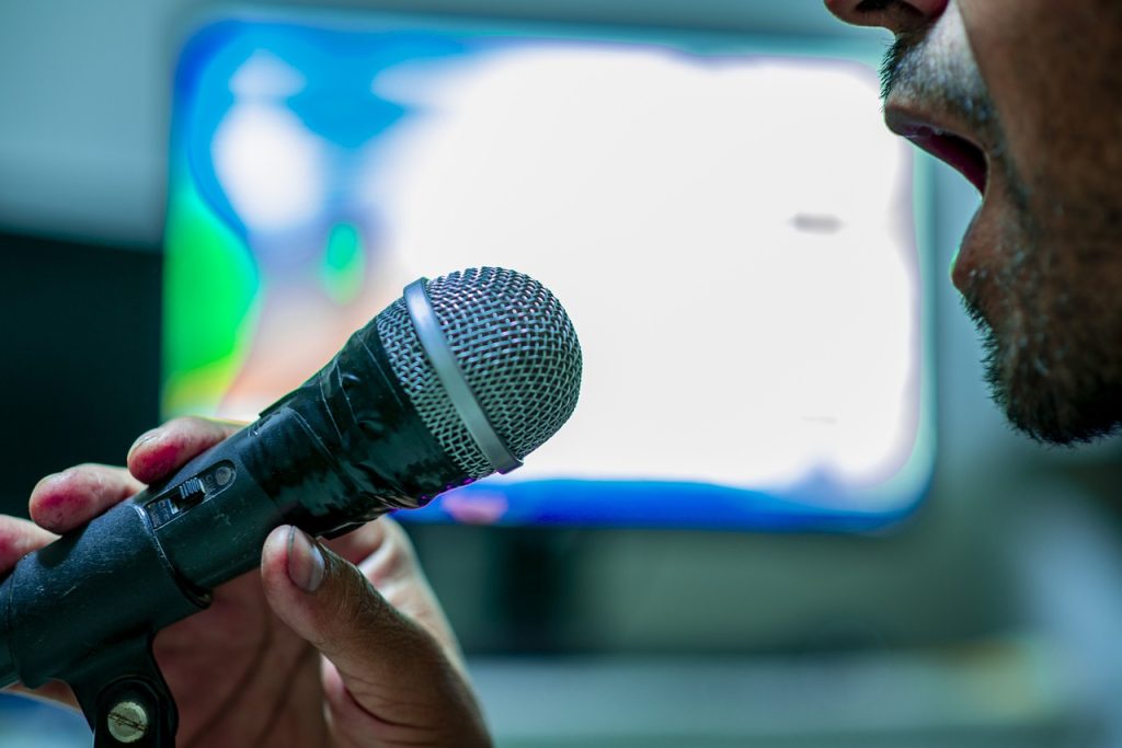 A man singing into a microphone with a karaoke screen behind him. We cannot see his whole face, just his open mouth