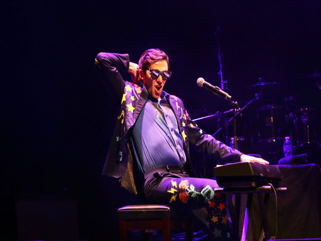 Tom Cridland sitting at a piano. He has one arm bent and holding his head. He is wearing sunsglasses and a star-studded jacket. He is pressing keys on a piano with his other hand and there is a microphone visible