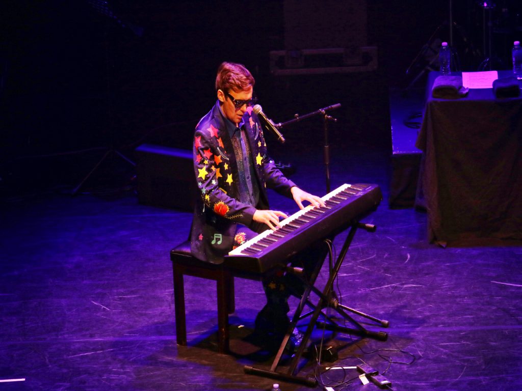 Tom Crudland playing the piano as part of his Elton John tribute. He is seated and in the middle of the image. He is wearing a star-studded jacket and has both hands on the keyboard 