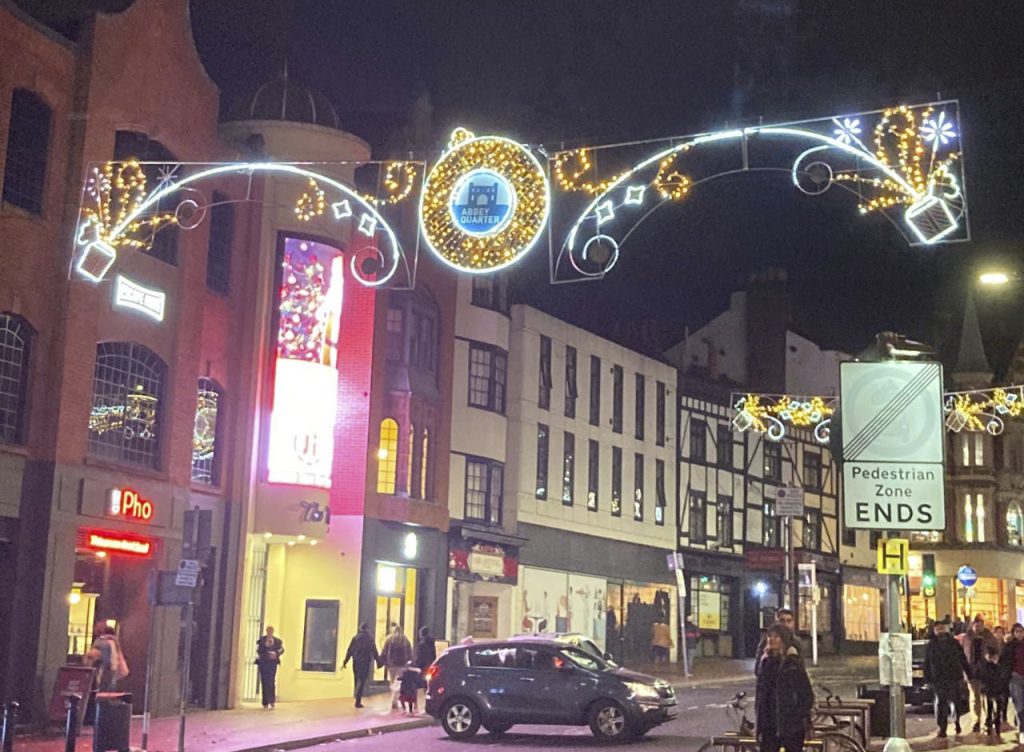 One of the new light displays in Reading town centre. This one is on King Street. The lights are on and in the middle is a circle featuring the image of Reading Abbey. Underneath the lights is a typical street scene with a car attempting a u-turn, and people walking along. 