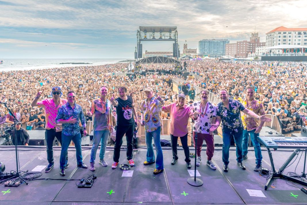 The Beach Boys on stage at a concert, watched by thousands of fans in the background. There are blue skies and cloud in the top of the image