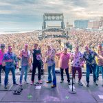 The Beach Boys on stage at a concert, watched by thousands of fans in the background. There are blue skies and cloud in the top of the image