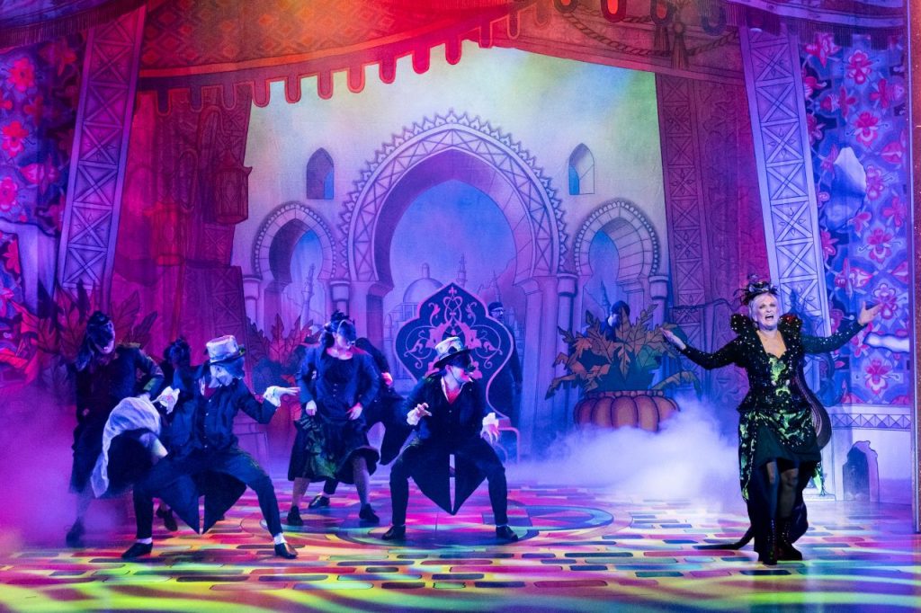 A scene from Dick Whittington at Theatre Royal Windsor set in Queen Rat's domain. Queen Rat is in on the right with her arms outstretched while the human rats are hunched up on the left. In the background is a kind of castle construction