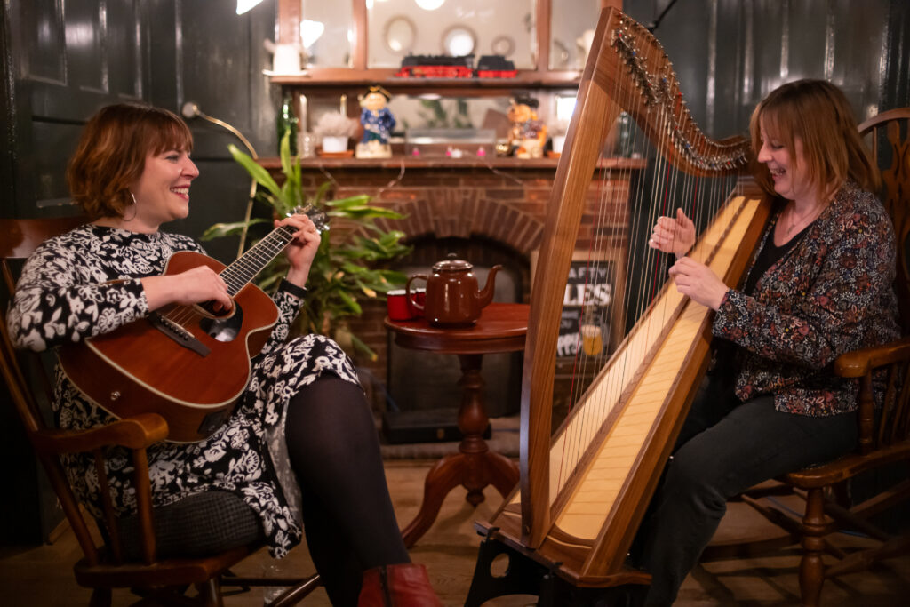 The Claras sitting down playing instruments: an acoustic guitar and a harp. In the background there is a small table with a teapot on there