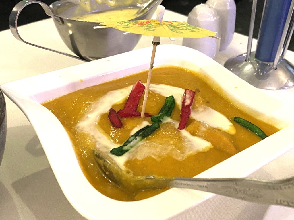 A bowl of chicken korma with a spoon in it. There is a yellow cocktail umbrella sticking out of it