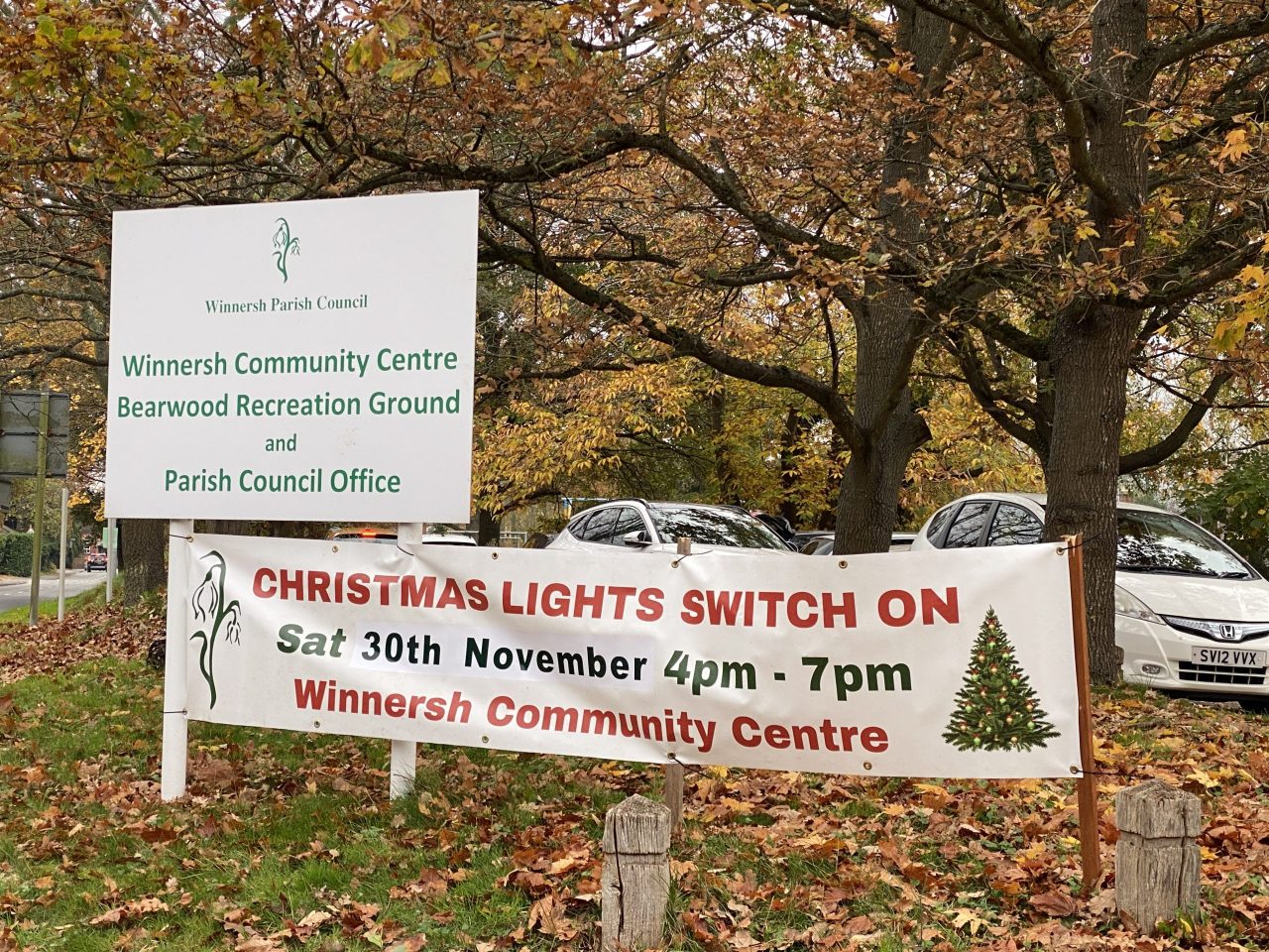 A banner promoting the launch of Winnersh Christmas lights, next to a sign at the entrance to Bearwood Recreation Ground. Behind the signs are tears shedding their leaves, which have turned orange and fallen on the ground. The banner reads: "Christmas light switch on, Sat 30th November 4pm - 7pm, Winnersh Community Centre'. The sign reads 'Winnersh Community Centre Bearwood Recreation Ground, and Parish Council Office'