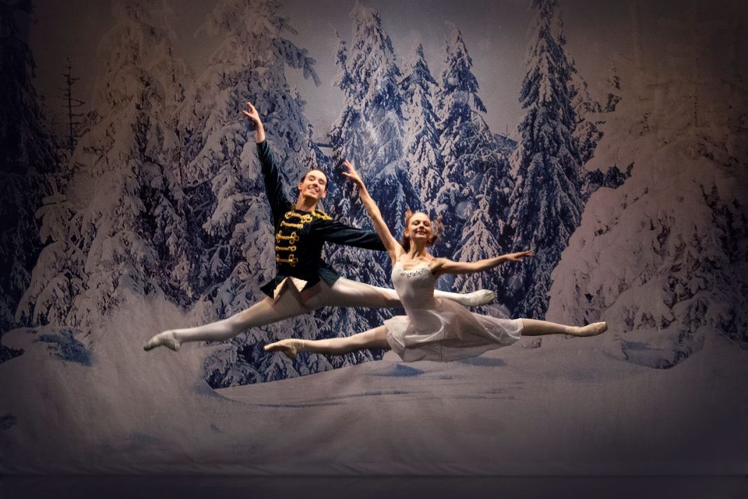 The prince and Clara leap for joy in a scene from The Nutcracker. They both have their legs outstretched and appear to be floating in the snowy woodland. There are alpine trees behind them, covered in snow