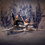 The prince and Clara leap for joy in a scene from The Nutcracker. They both have their legs outstretched and appear to be floating in the snowy woodland. There are alpine trees behind them, covered in snow