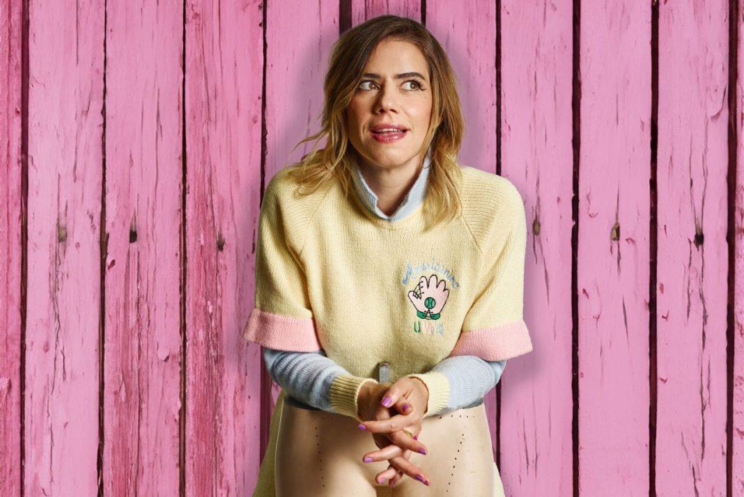 Comedian Lou Sanders possing. We can see her head and upper body. She is wearing a yellow jumper and holding the legs of a shop dummy in her hands. She is standing against a wood-panelled background. The wood has been stained pink