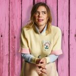 Comedian Lou Sanders posing. We can see her head and upper body. She is wearing a yellow jumper and holding the legs of a shop dummy in her hands. She is standing against a wood-panelled background. The wood has been stained pink