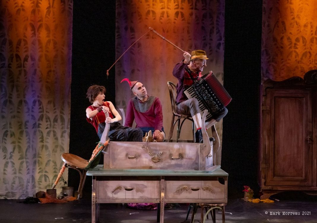 A production shot from the Norden Farm production of Pinocchio shows the three cast members in a scene. They appear to be sitting on a chest of drawers. One of the cast is holding a chair, another has a whip and an accordian. The third, in the middle, is wearing a red, wavy hat of some kind.