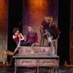 A production shot from the Norden Farm production of Pinocchio shows the three cast members in a scene. They appear to be sitting on a chest of drawers. One of the cast is holding a chair, another has a whip and an accordian. The third, in the middle, is wearing a red, wavy hat of some kind.