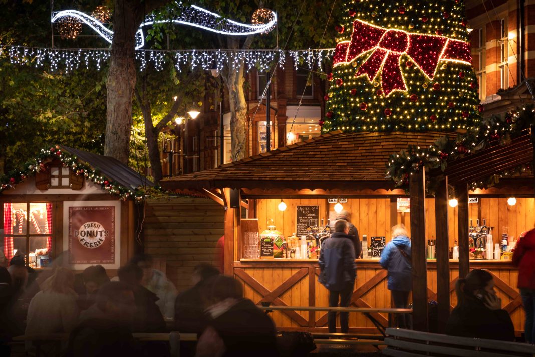 A wooden hut as part of Reading's Christmas market. The picture is taken at night so the hut is lit up. Staff inside are serving two customers. Behind the hut it is possible to see Christmas lights and a Christmas tree.