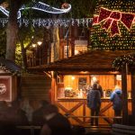 A wooden hut as part of Reading's Christmas market. The picture is taken at night so the hut is lit up. Staff inside are serving two customers. Behind the hut it is possible to see Christmas lights and a Christmas tree.