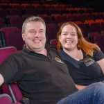 Steve and Sarah Boden are sitting in theatre seats and smiling. There are rows of red velvet seats behind them