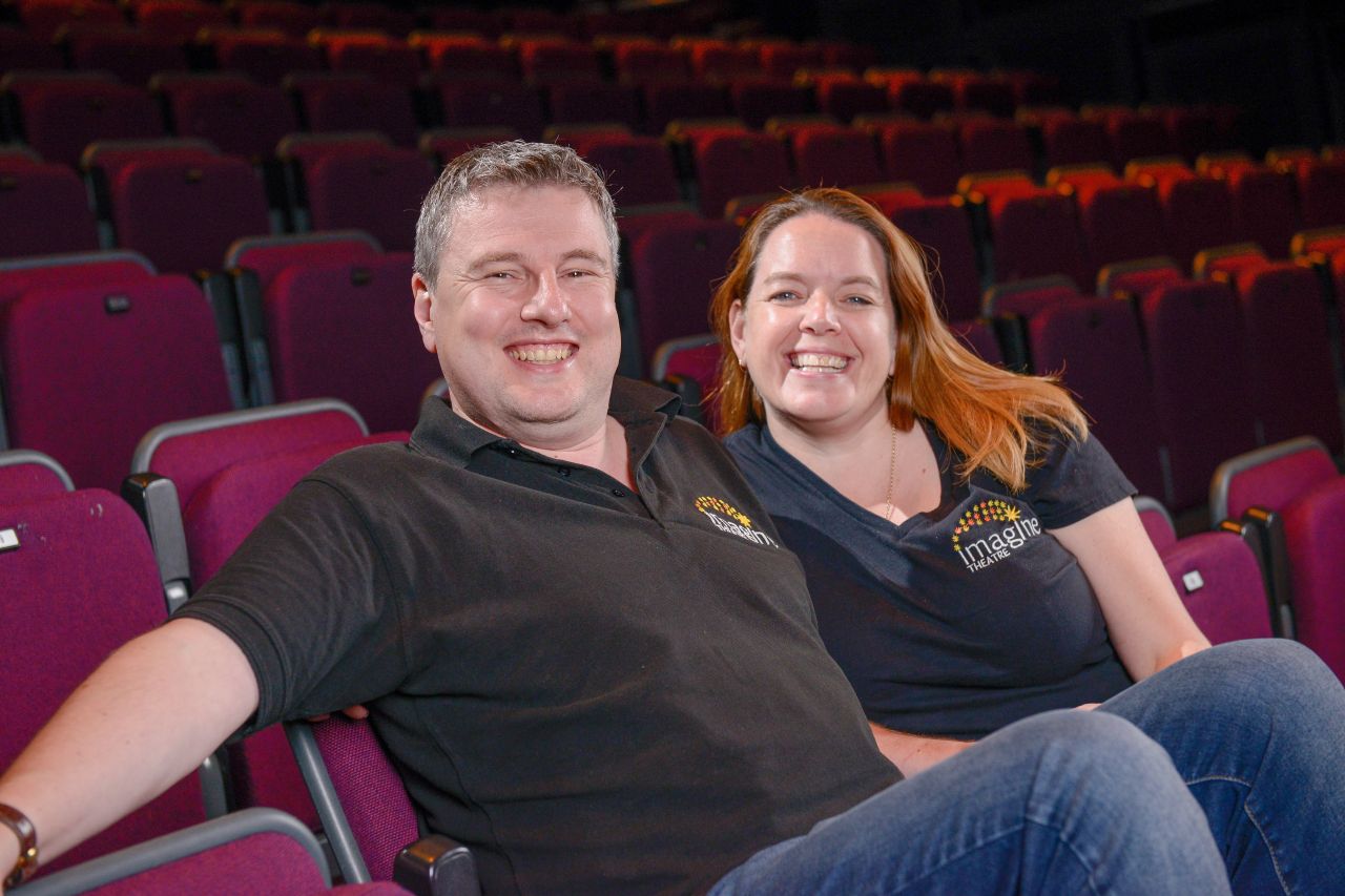 Steve and Sarah Boden are sitting in theatre seats and smiling. There are rows of red velvet seats behind them