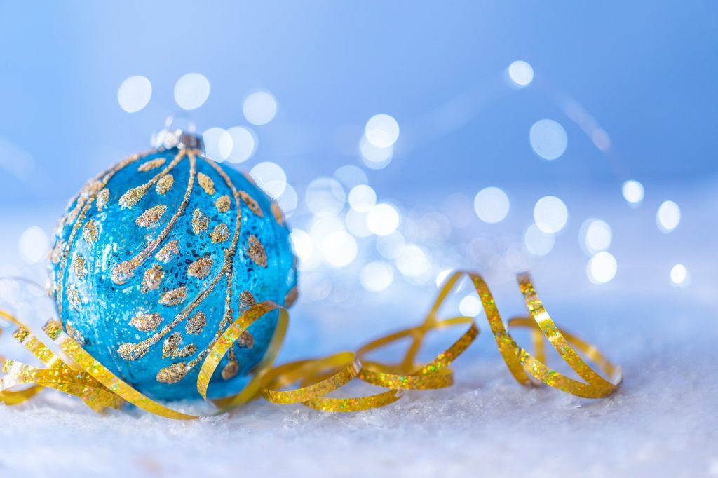 A blue Christmas bauble sitting on a golden ribbon on a snowy background with some white festive lights in the background