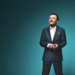 Chris McCausland on the right of the image looking up. We see him from his knees up and he is wearing a suit.