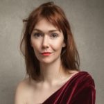 Elf Lyons head and shoulders image. She is wearing a red velvet dress on one shoulder, and has a naked neckline on the other.