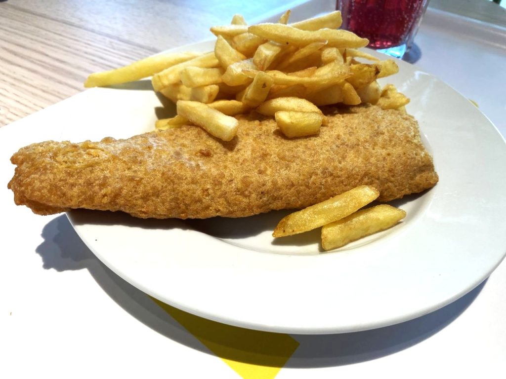 A plate of fish and chips on a tray with a red coloured drink behind the plate