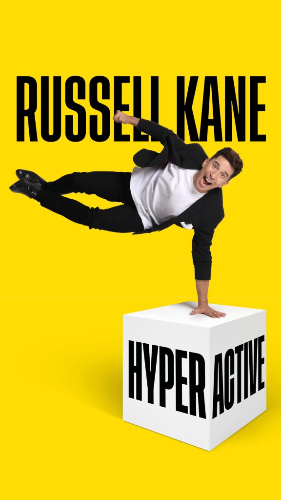 Russell Kane doing a one hand stand on a white cube