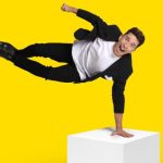Russell Kane doing a one hand stand on a white cube