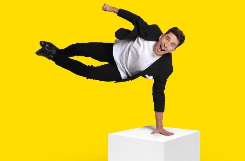 Russell Kane doing a one hand stand on a white cube