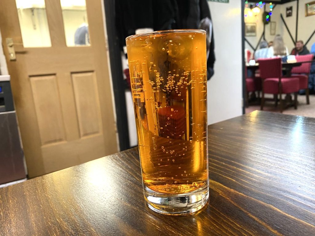 Irn Bru is one of the many soft drinks available at Sweeney and Todd's in Reading. A glass of it is on a brown stained table. On the left is a door leading into the kitchen, and on the right, blurred and in the distance, are some of the tables.