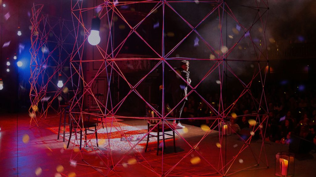 James Phelan on a stage with a microphone. He is on the right and is msall. Our view is obscured by a grid in front of the camera