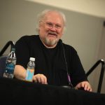 Sixth Doctor Who Colin Baker sitting at a desk with two microphones and a bottle of water next to him. He is appearing at Reading Comic Con