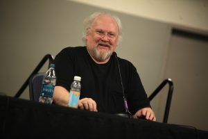 Sixth Doctor Who Colin Baker sitting at a desk with two microphones and a bottle of water next to him. He is appearing at Reading Comic Con