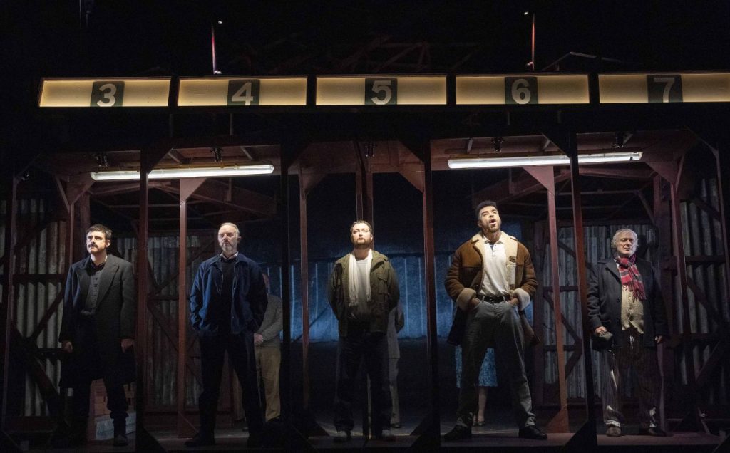 Five men on stage under numbered booths in a scene from Boys From The Blackstuff