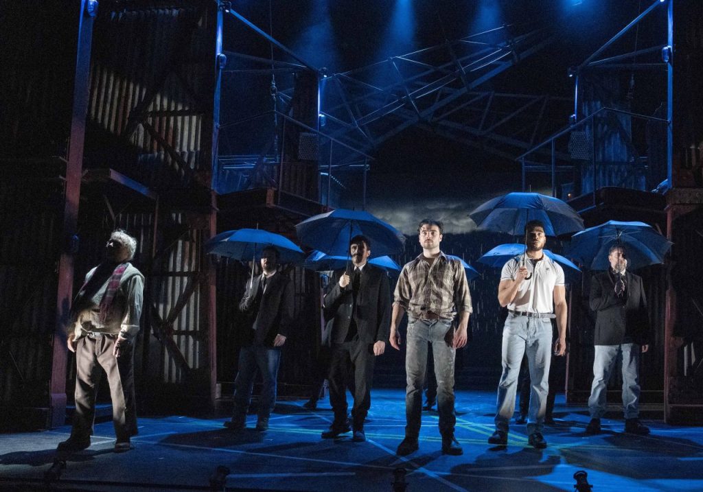 The cast of Boys From The Blackstuff holding umbrellas on a darkened stage