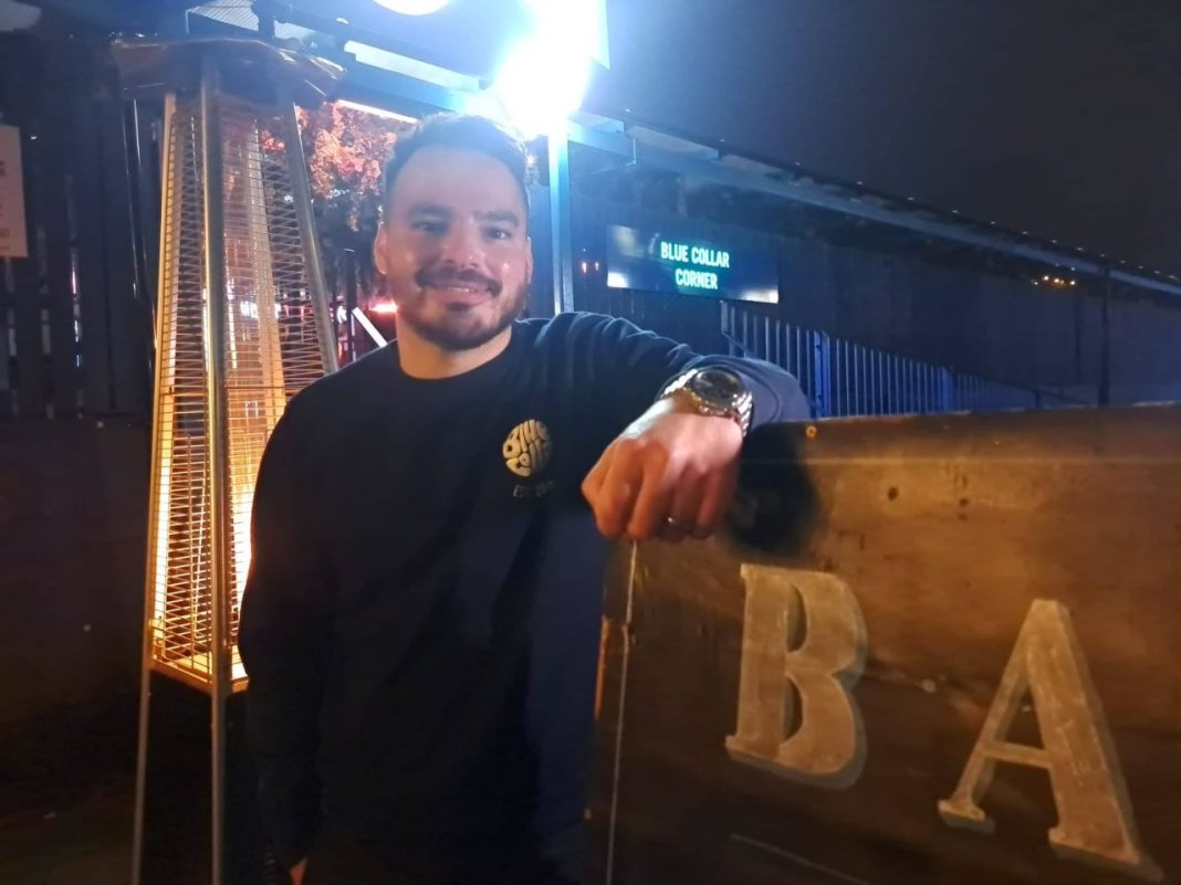 Glen Dinning from Blue Collar Corner leaning on a sign that says BAR - only the first two letters are visible