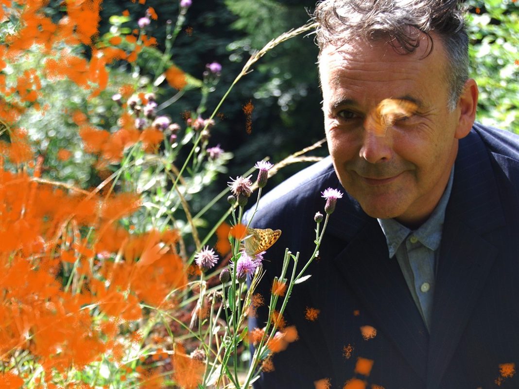 Rob Newman on the right of the picture. His face is partially obscured by a butterfly. He is outdoors and next to some flowers - probably poppies