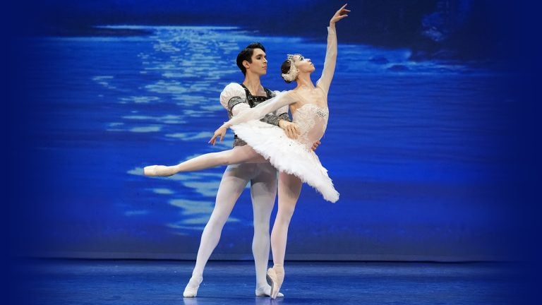 Varna International is presenting Swan Lake, a ballet. Pictured are a male and female ballet dancer on stage with a blue background