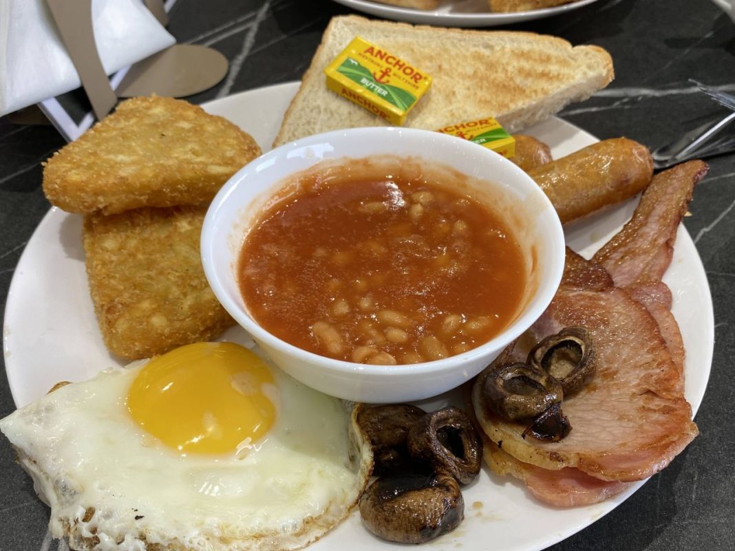 A full english at Woodley Café. Clockwise from bottom left: a fried egg, two hash browns, toast with butter, sausages, bacon, and mushrooms. In the middle is a bowl of baked beans