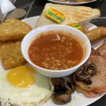 A full english at Woodley Café. Clockwise from bottom left: a fried egg, two hash browns, toast with butter, sausages, bacon, and mushrooms. In the middle is a bowl of baked beans