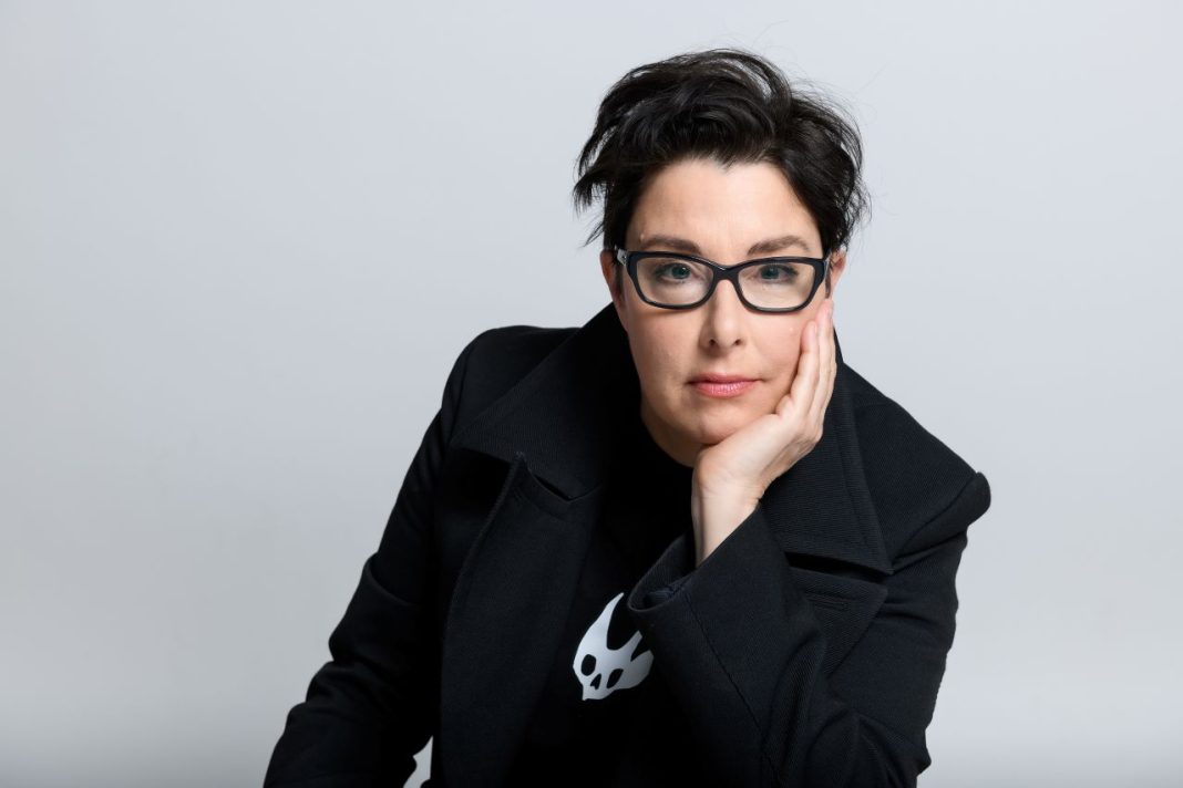 Sue Perkins with one hand resting on her face. She is wearing glasses and looking at us