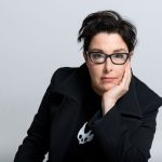 Sue Perkins with one hand resting on her face. She is wearing glasses and looking at us