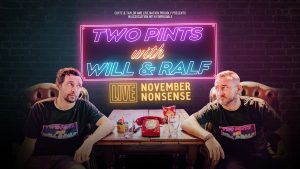 Ralf Little and Will Mellor sitting either side of a desk with a sign behind them that reads Two Pints with Wll and Ralf LIVE November Nonsense. This is a neon effect on the desk between them are two empty pints, an old rotary dial telephone that is red and a packet of crisps.