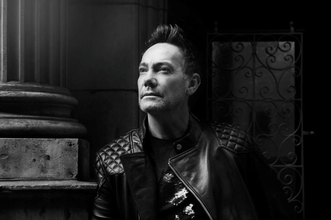 Craig Revel Horwood head and shoulders. This is a black and white image and Craig is in the middle of the picture. He is wearing a leather jacket and standing next to a column, looking upwards