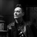 Craig Revel Horwood head and shoulders. This is a black and white image and Craig is in the middle of the picture. He is wearing a leather jacket and standing next to a column, looking upwards