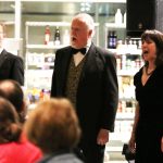 The Opera D'Amici singers lined up. Their mouths are open because they are singing. They are wearing dinner jackets and evening wear. In the foreground are the backs of people's head watching the singers.