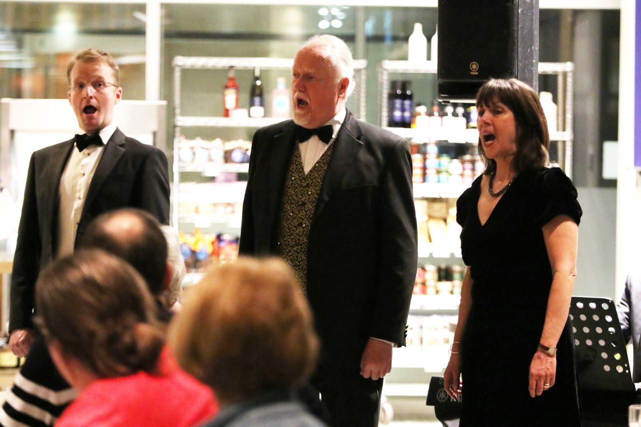 The Opera D'Amici singers lined up. Their mouths are open because they are singing. They are wearing dinner jackets and evening wear. In the foreground are the backs of people's head watching the singers.
